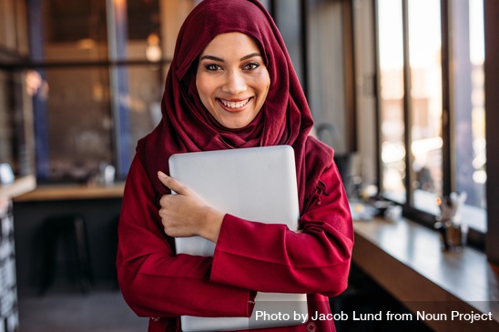 A women in a hijab smiles to the camera and clutches a laptop